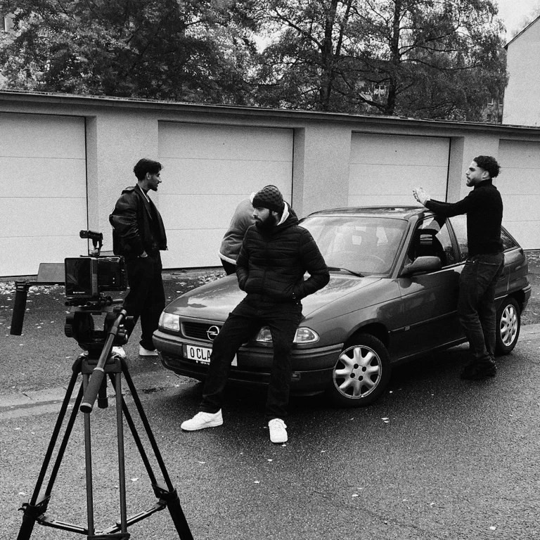 3 junge Männer werden von einer Kamera auf dem Stativ gefilmt, wie sie an einem Auto lehnen